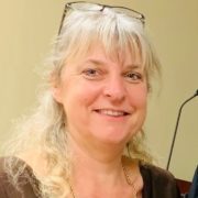 Photo of a woman with light hair, bangs, glasses on the top of her head, and a dark brown shirt.