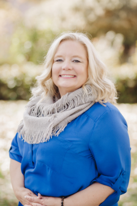 Photo of woman with light blonde hair in a cobalt blue shirt and gray scarf, standing outside.