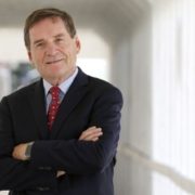A photo of a man with short brown hair wearing a black suit with a light blue shirt and dark red tie with arms crossed, standing in a hallway.