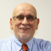 A man who is bald and had a white beard. He is wearing a light blue collared shirt with a reddish-orange tie and glasses.