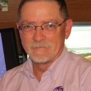 A photo of a man with dark grey hair and white/gray beard wearing a light pink shirt and glasses.