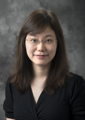 A photo of a woman with long brown hair and bangs wearing a black suit jacket in front of a gray background.