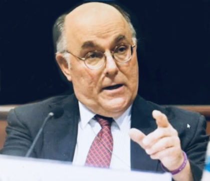 A man with black and gray hair and glasses wearing a suit, sitting in front of a microphone and pointing his index finger while talking.