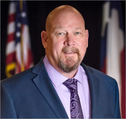 A photo of a man standing in front of an American flag. He is bald and has a medium-brown beard.