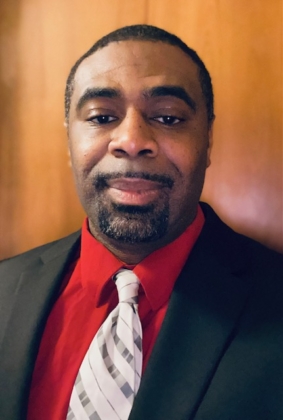 A photo of a man with dark brown hair and beard wearing a red shirt, white tie and black suit jacket in front of a wood paneled wall.