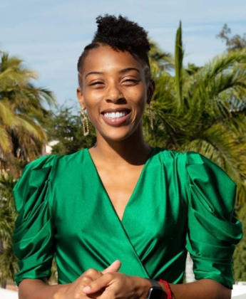 Photo of woman with dark brown hair in a green shirt with palm trees in the background.