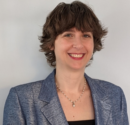 A photo of a woman with short brown hair wearing a black shirt with a blue/gray suit jacket in front of a light gray background.