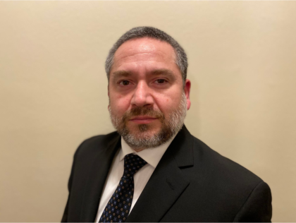 A photo of a man with dark brown and gray hair and beard wearing a black suit in front of a beige background.