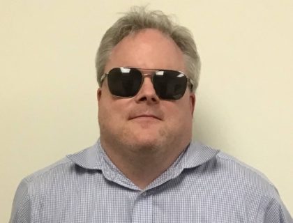 Man in a light blue shirt with sunglasses in from of an off-white background.